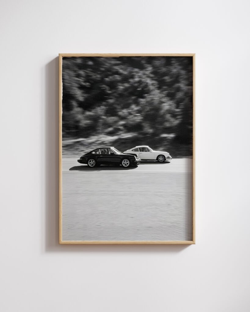 Framed black-and-white photo of two vintage Porsche 911s racing side by side, blurred trees behind.