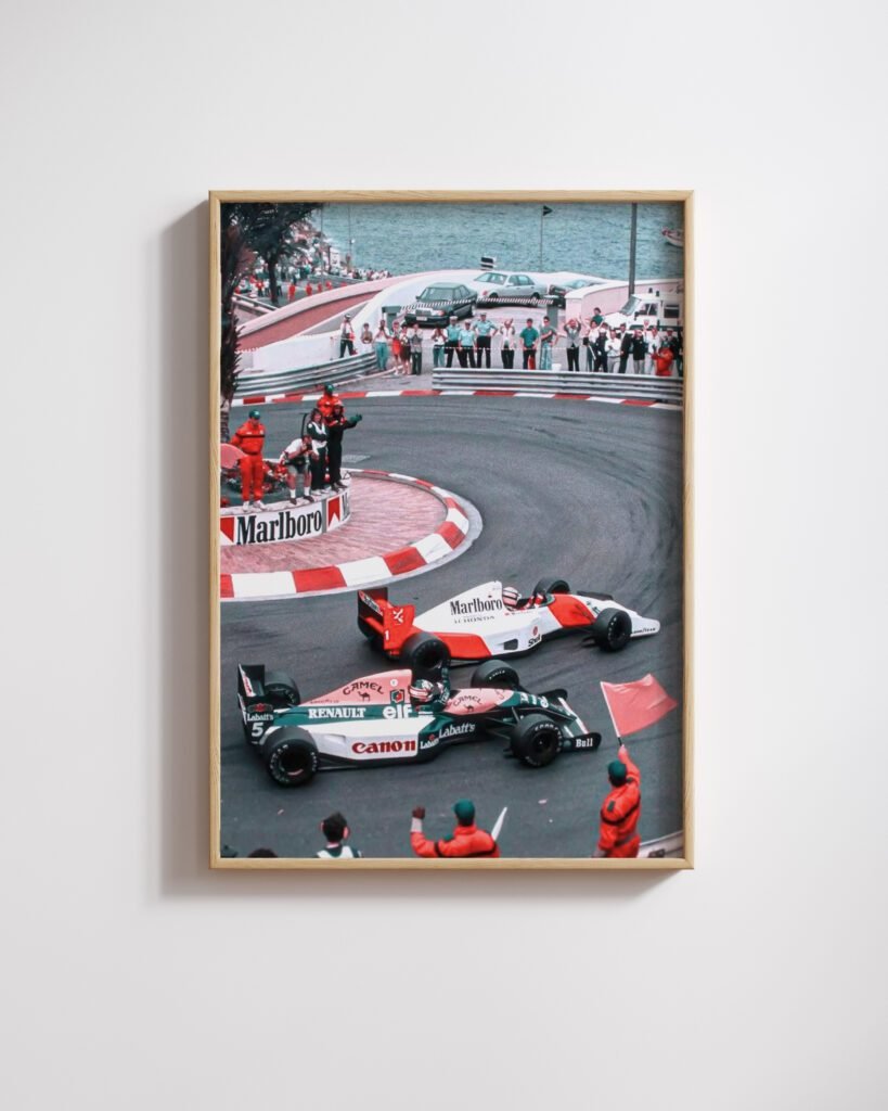 Framed vintage colour photo of two open-wheel race cars cornering past spectators, on a white wall.