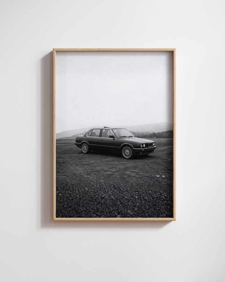Framed black-and-white photo of a black BMW E30 sedan parked on a gravel lot in misty hills.
