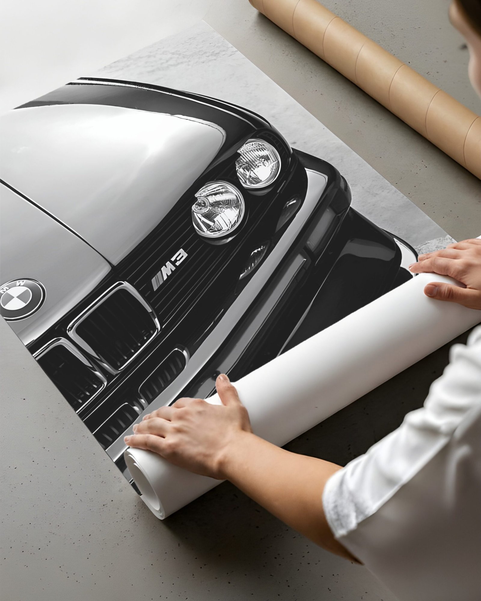 A person rolls up a black-and-white BMW M3 print beside a cardboard shipping tube.