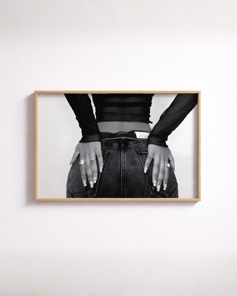 Framed black-and-white close-up of a woman's hands resting on jean back pockets, wearing a sheer top.