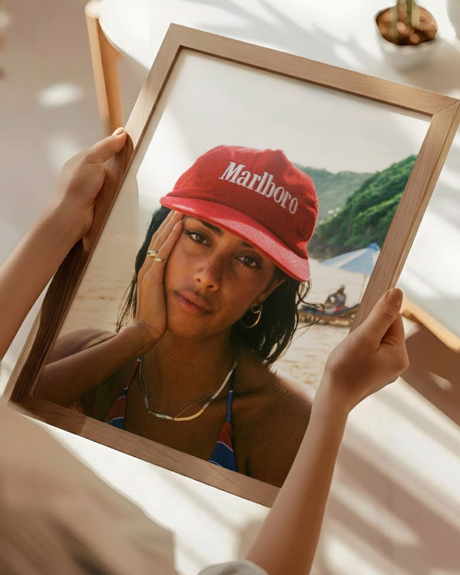 Hands holding a framed beach portrait of a woman in a red branded cap above a café table.