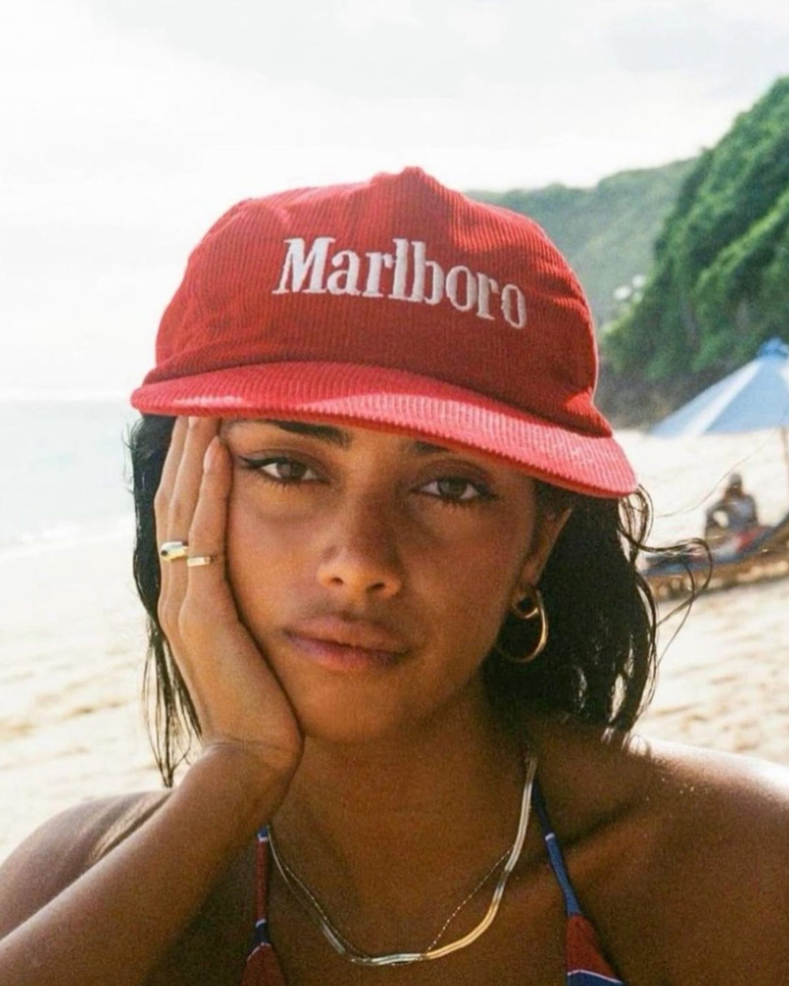 Close-up beach portrait of a woman in a red branded corduroy cap, resting her cheek on her hand.