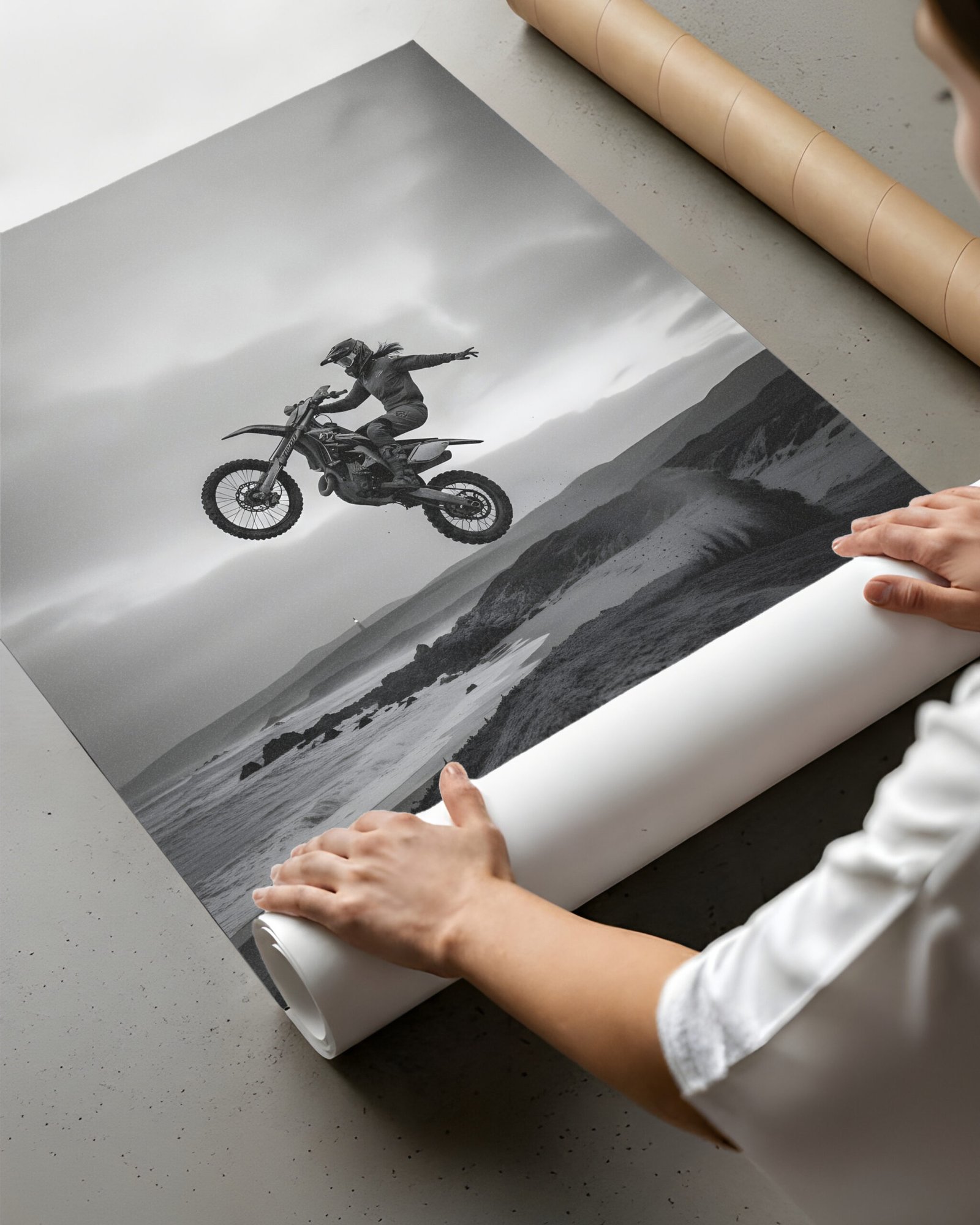 A person unrolls a black-and-white motocross jump poster print beside a cardboard shipping tube.