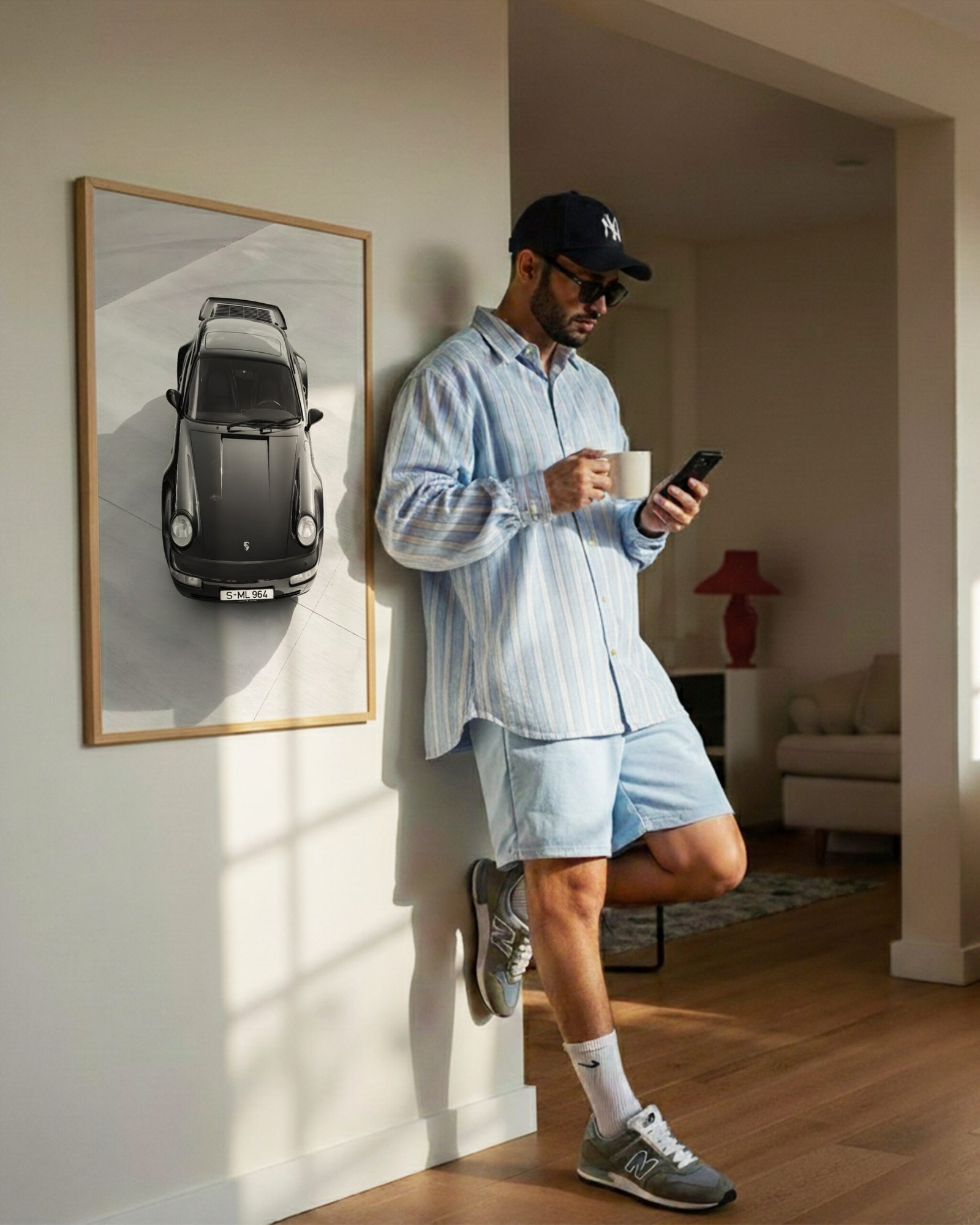 Framed black-and-white Porsche print on a wall beside a man in a cap holding coffee and a phone.