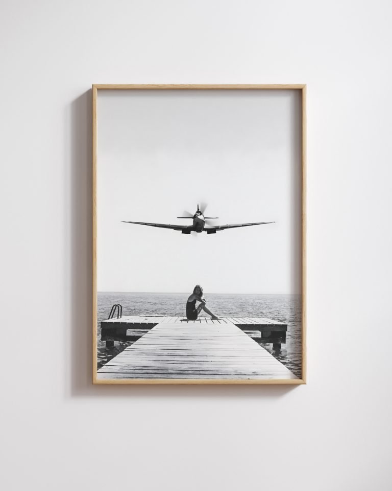 Framed black-and-white print of a vintage propeller plane flying low over a woman seated on a dock.