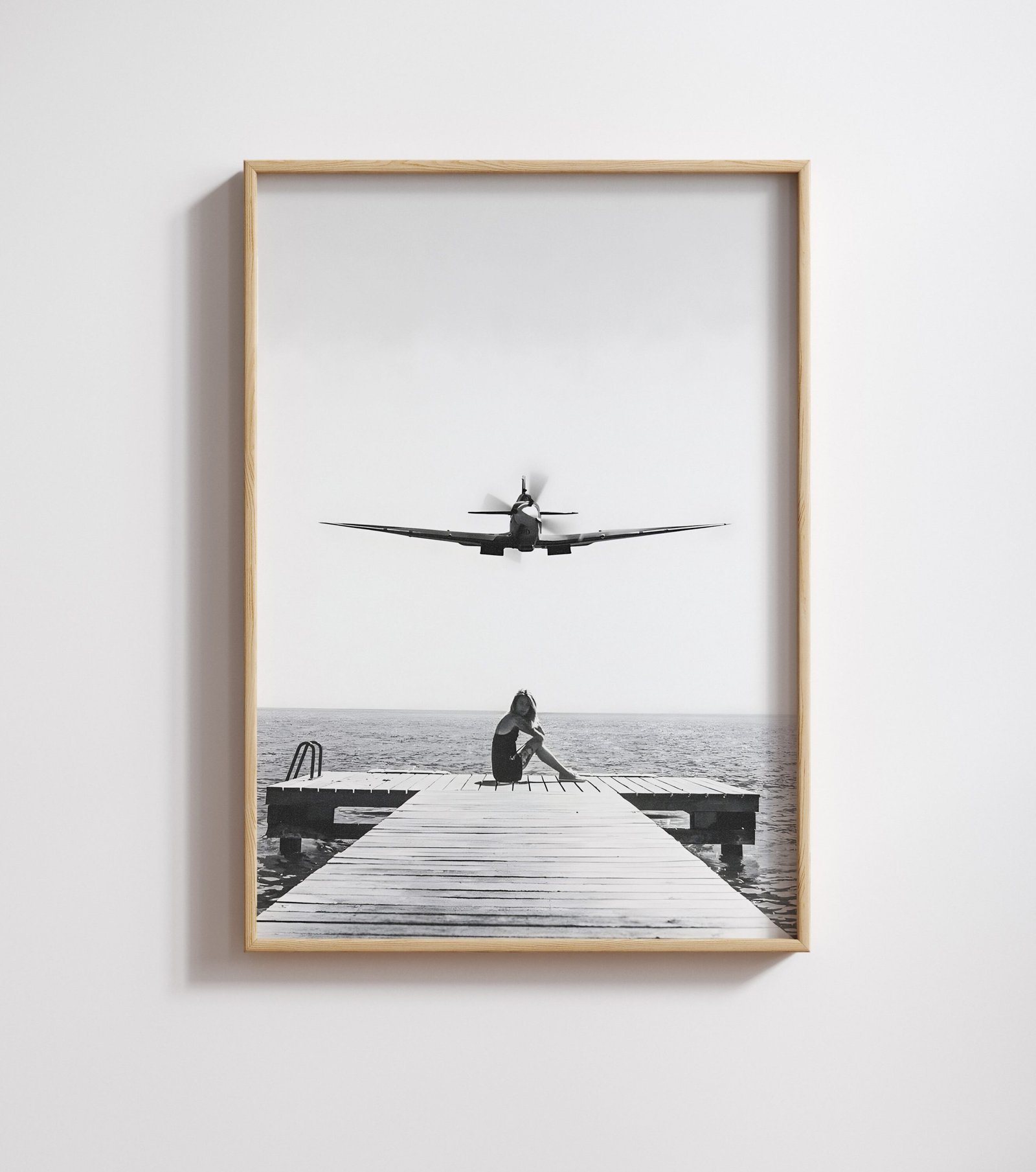 Framed black-and-white print of a vintage propeller plane flying low over a woman seated on a dock.