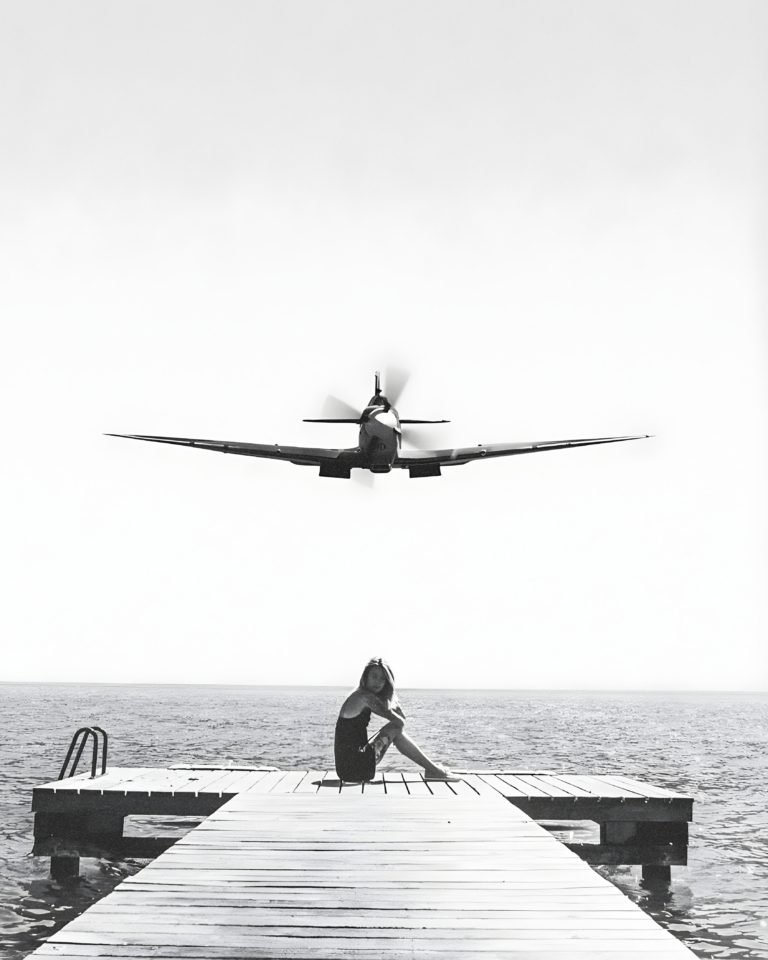 Black-and-white photo of a vintage propeller plane flying low above a woman seated on a dock.