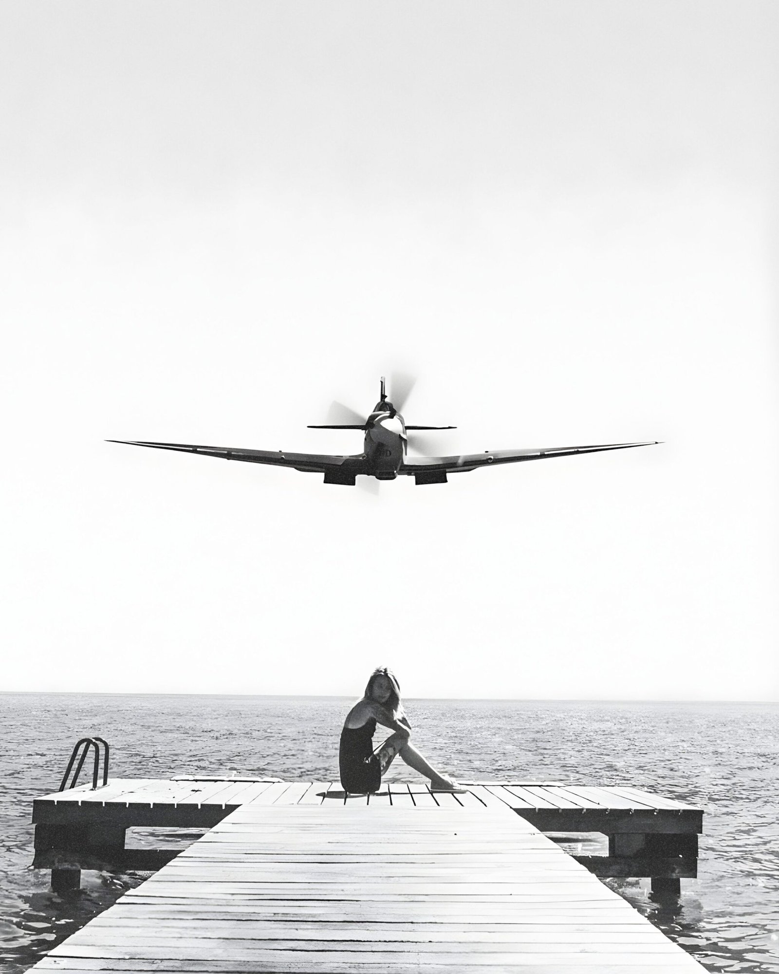 Black-and-white photo of a vintage propeller plane flying low above a woman seated on a dock.
