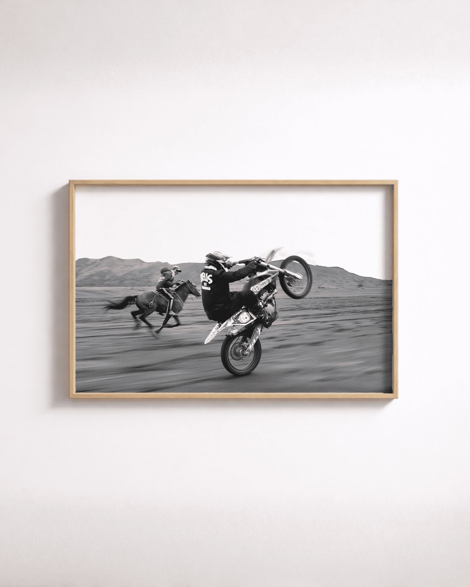 Framed black-and-white photo of a motocross rider popping a wheelie ahead of a galloping horse and rider in the desert.